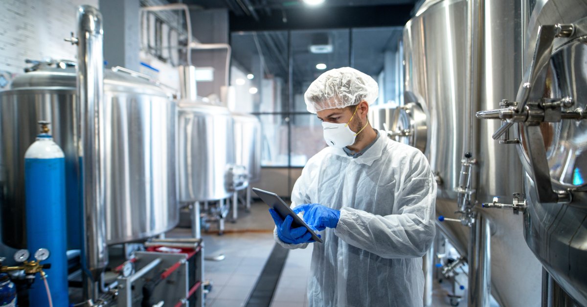 A man wearing blue gloves and a facemask, holding a clipboard. There is silvery machinery all around him.