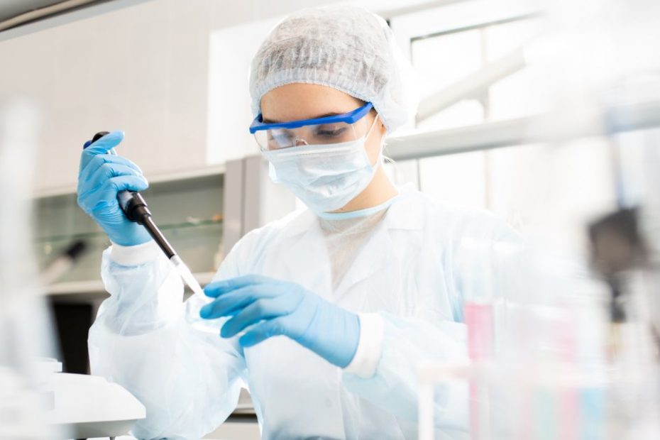 A health-care professional wearing protective glasses and a facemask, holding a black object in her right hand.