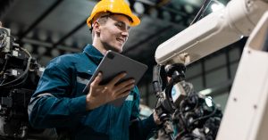 A smiling man who is wearing a yellow hard hat and holding a tablet in his hand is inspecting a mechanical arm.