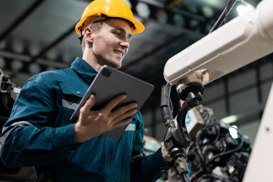 A smiling man who is wearing a yellow hard hat and holding a tablet in his hand is inspecting a mechanical arm.