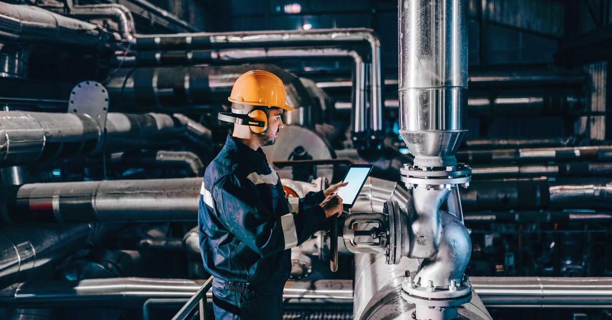 A man wearing a hard hat and ear protection is holding a tablet in one hand as he stands in front of a large pipe.