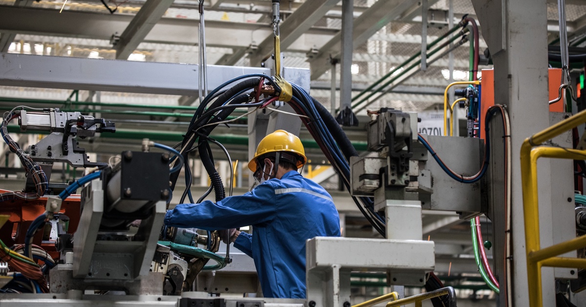 A person in a hard hat and a blue jumpsuit is bent over some heavy machinery and is working on it with his hands.
