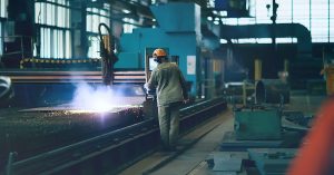 A man wearing protective equipment stands next to a piece of machinery. The machine is creating sparks.