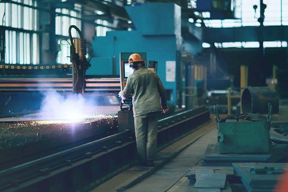 A man wearing protective equipment stands next to a piece of machinery. The machine is creating sparks.
