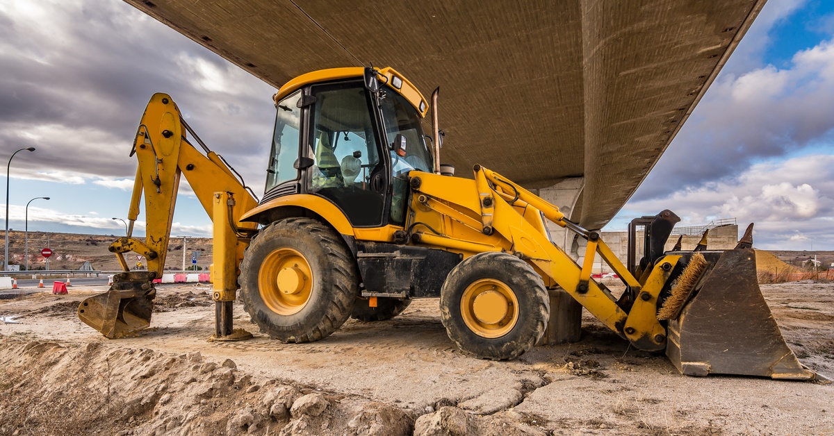 A yellow backhoe is under a bridge; there is no one inside the backhoe. The ground is dirt and there are clouds in the sky.