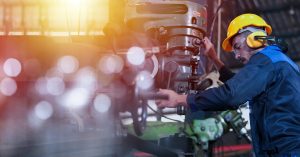 A man in a blue jumpsuit and a hard hat is holding onto a large piece of manufacturing machinery.