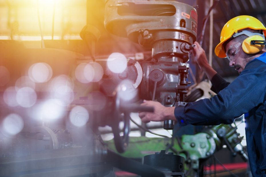 A man in a blue jumpsuit and a hard hat is holding onto a large piece of manufacturing machinery.