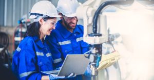 Two engineers are standing next to each other in a factory workshop. Both are wearing blue jumpsuits and hard hats.
