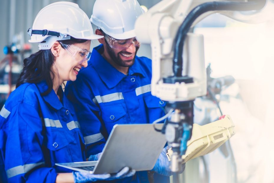 Two engineers are standing next to each other in a factory workshop. Both are wearing blue jumpsuits and hard hats.