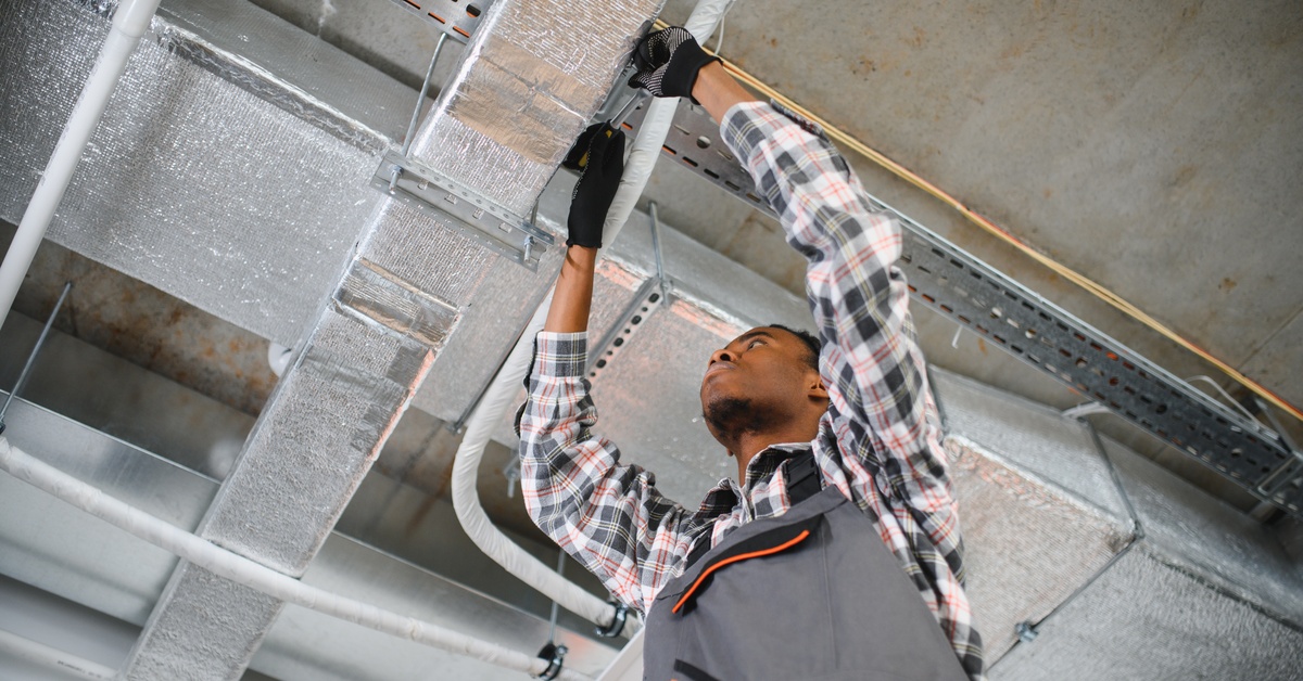 A man wearing a flannel shirt and protective gloves is reaching both hands up to a ventilation shaft.