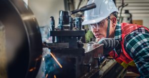 A man with a protective helmet and glasses is looking closely at a machine. He’s wearing an orange vest.