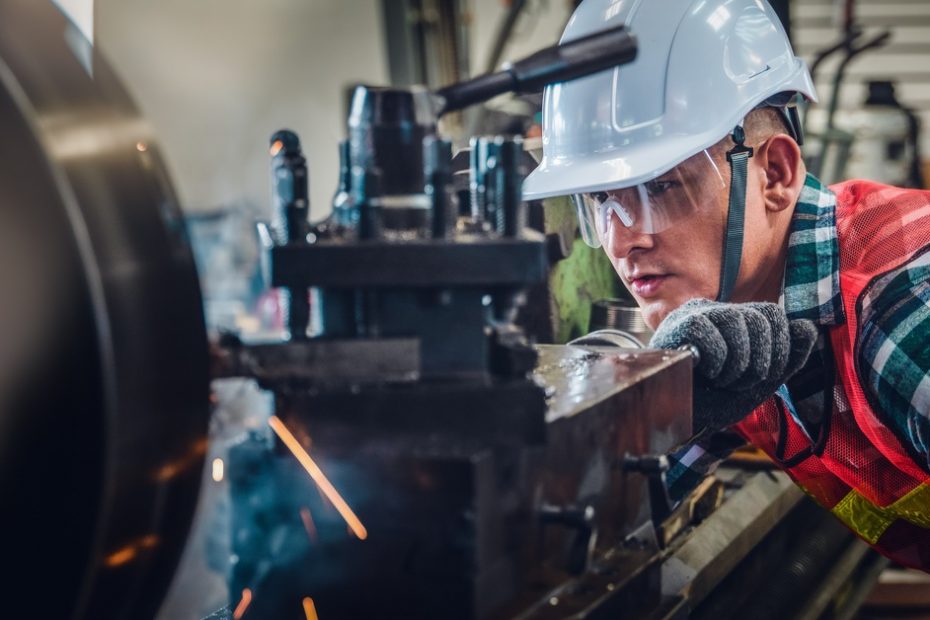 A man with a protective helmet and glasses is looking closely at a machine. He’s wearing an orange vest.