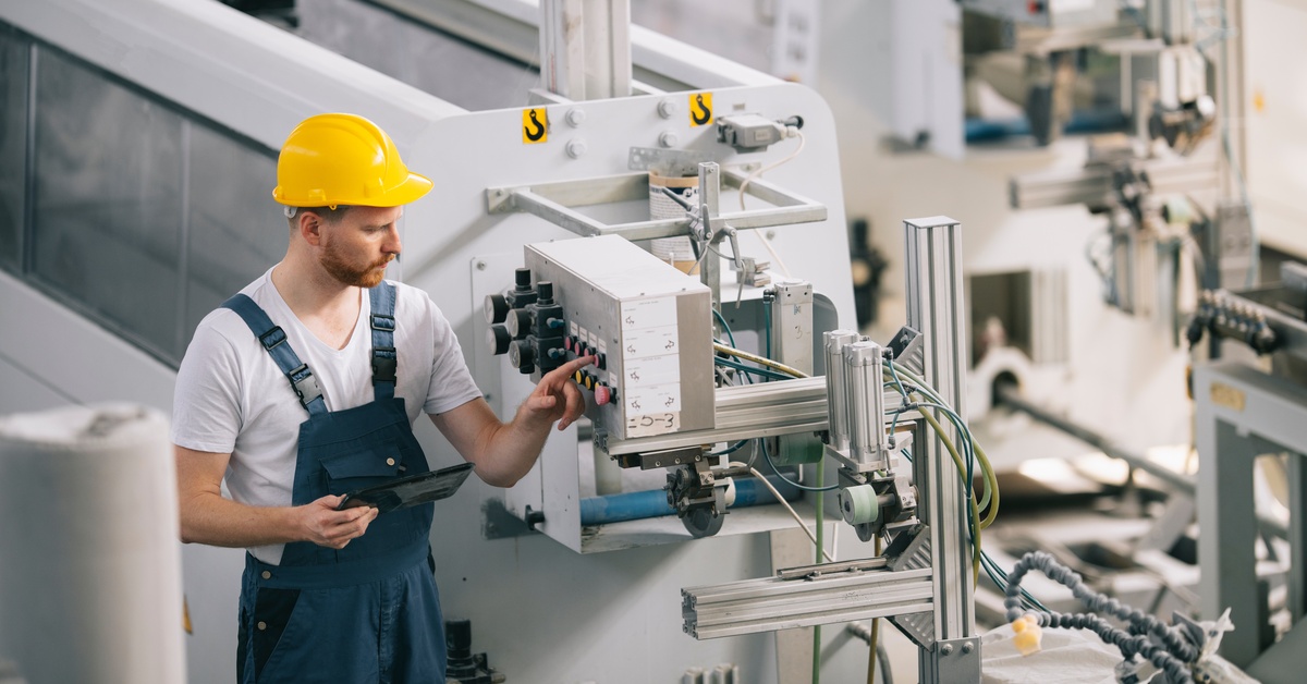 A man in a yellow hard hat and blue overalls is looking at a machine. He’s holding a tablet and pointing at a button.