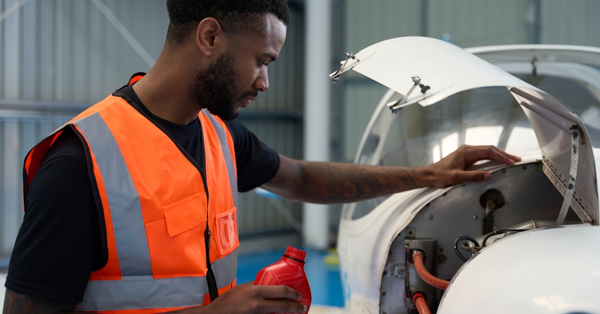 A man in an orange vest is looking at an open compartment on a plane. The compartment has mechanical parts in it