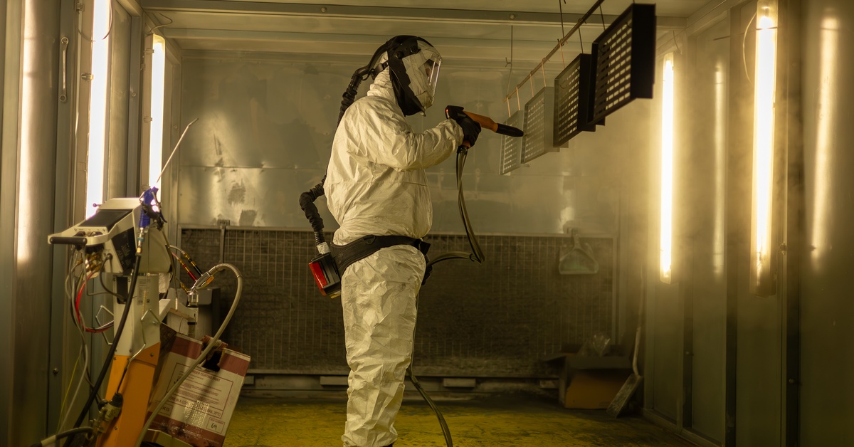 A man is using an applicator to spray several pieces of metal that are suspended above the floor.
