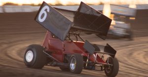 A red dirt race car is driving on a dirt track. There is a large spoiler on the roof of the race car.