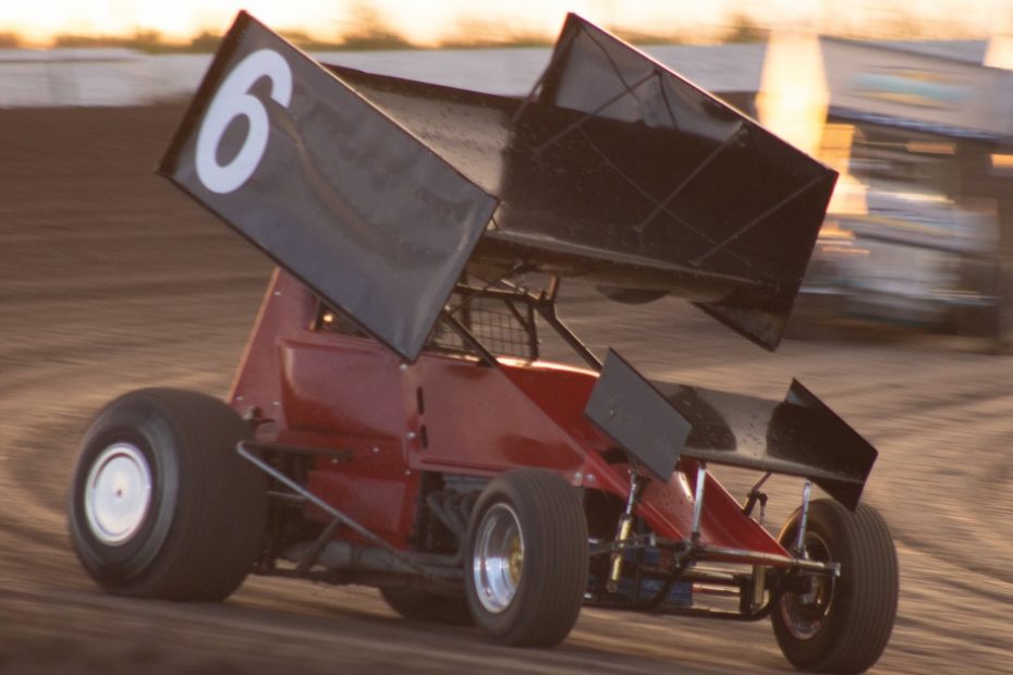 A red dirt race car is driving on a dirt track. There is a large spoiler on the roof of the race car.
