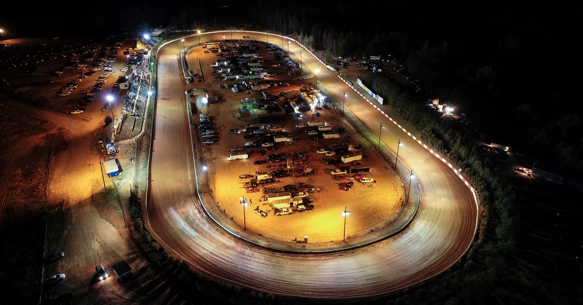 An aerial view of a racetrack at night. There are lights illuminating the race track and cars parked in the center of the track.