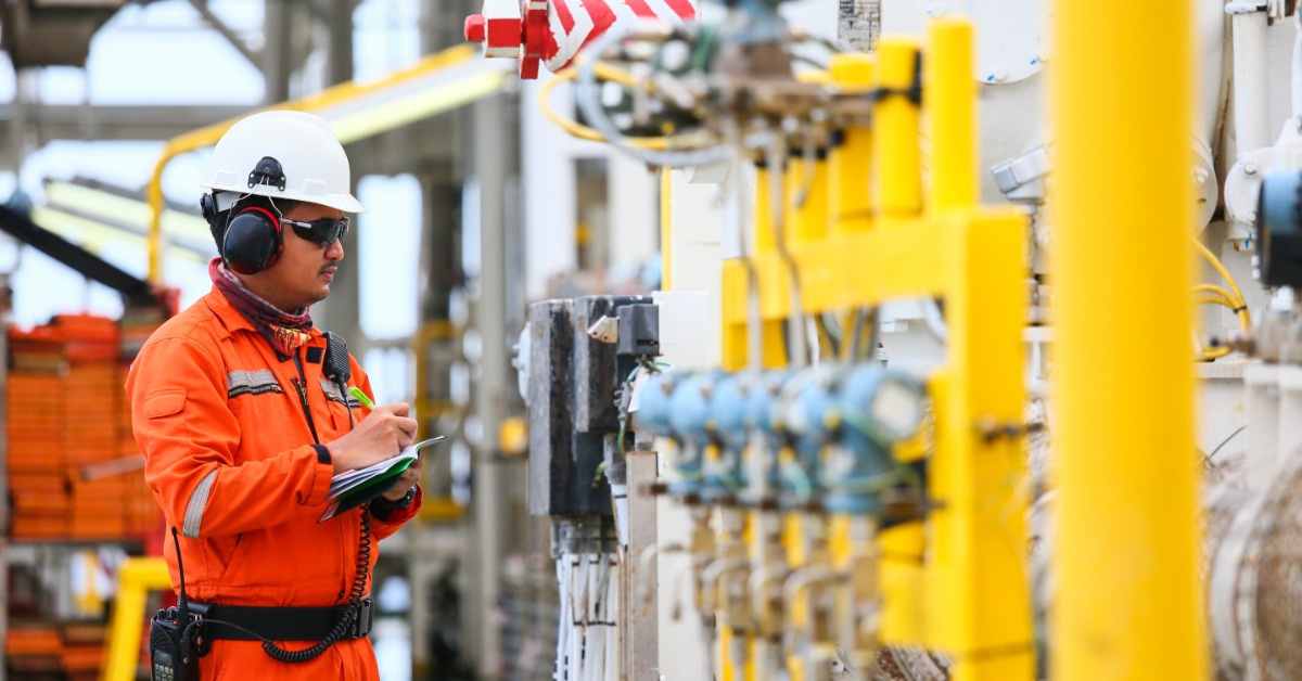A man is wearing ear protection and a hard hat as he looks at several metal objects. He is holding a clipboard.