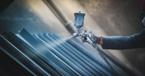 A person with a spray gun is applying a spray to a metal surface. The person is wearing gloves as they apply the coating.