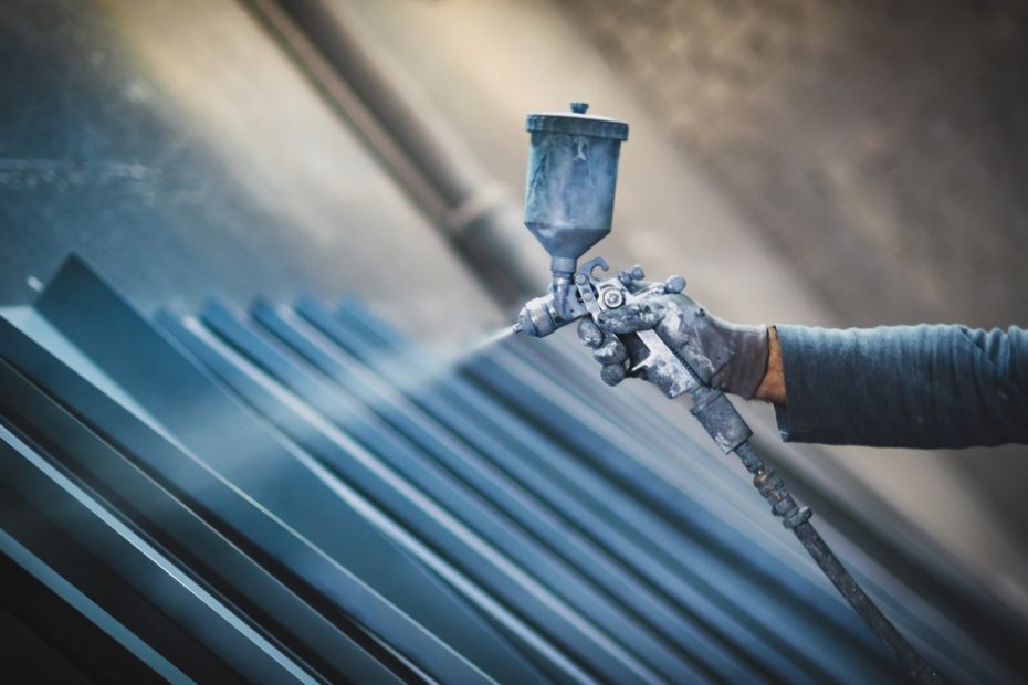 A person with a spray gun is applying a spray to a metal surface. The person is wearing gloves as they apply the coating.