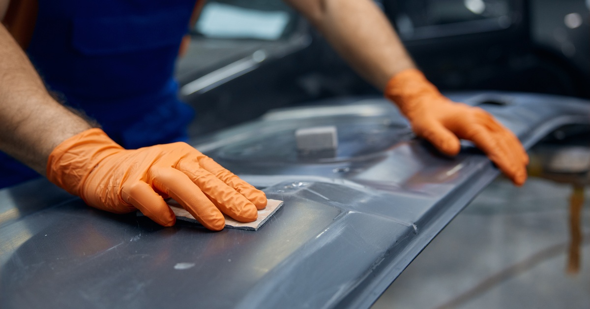 A man in a red shirt, blue overalls, and wearing gloves is placing their hands on a piece of metal.