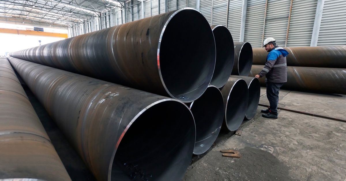 Long, metal pipes are stacked up and a man is standing next to the pipes. The pipes stretch far into the background.