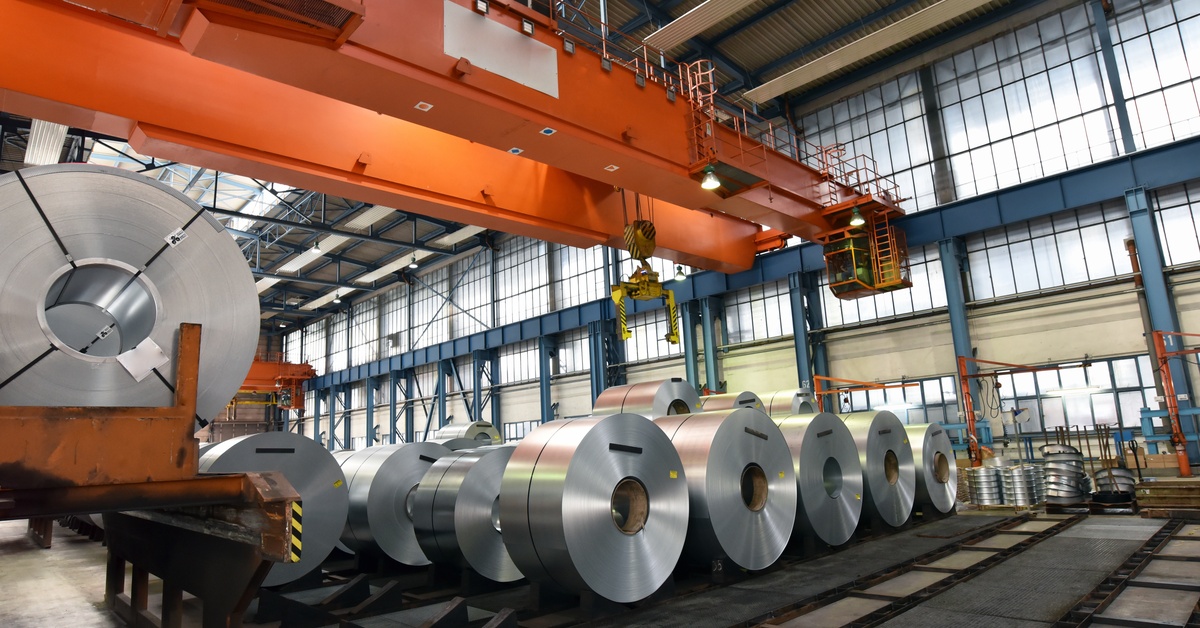 Metal cylindrical tubes are lines up on a factory floor. There is an orange crane above the tubes.