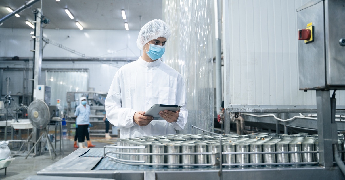 An industrial worker in personal protective gear is inspecting metal cans in a food processing plant.