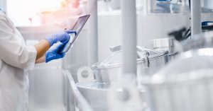 A factory worker wearing a white coat and blue gloves is inspecting a production line and holding a tablet.