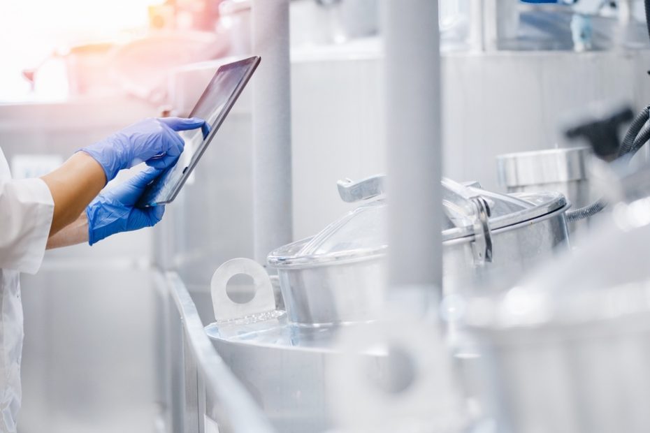 A factory worker wearing a white coat and blue gloves is inspecting a production line and holding a tablet.