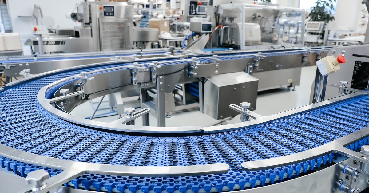 In an industrial production plant, there is a blue conveyor belt in the foreground with metal packaging equipment behind it.