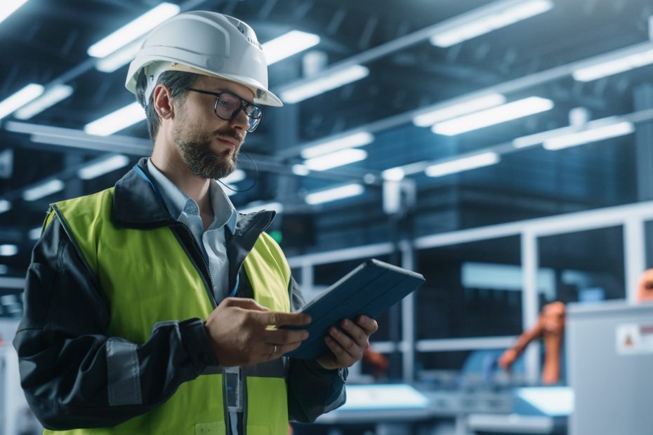 A man in a high-visibility vest and a hard hat is looking at a tablet in his hands. He’s in an industrial warehouse.