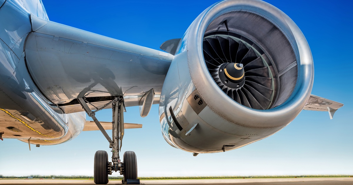 An airplane’s jet engine. The plane is on a tarmac and the sun is shining. There is foliage far in the background.