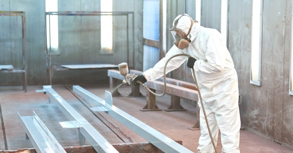A man in a white jumpsuit is using a hose to spray a large piece of metal with a coating.