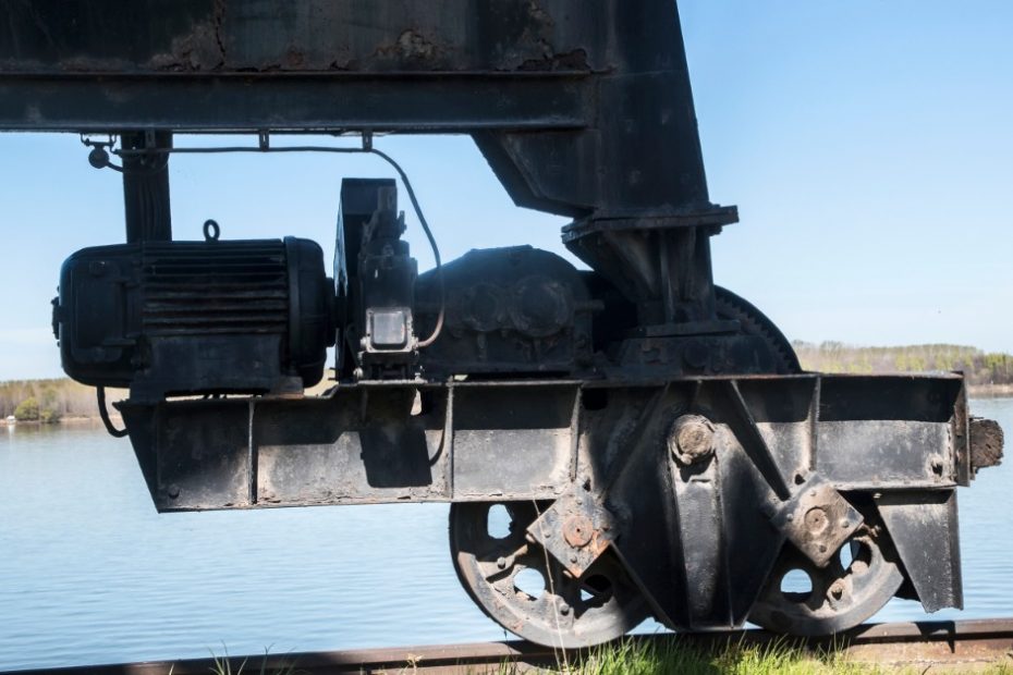 A metallic wheel is on a rail. There is grass growing next to the rail and the sky is blue in the background.