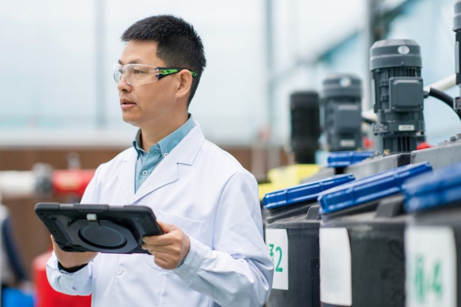 A man in a white lab coat is standing in the middle of an industrial setting. He’s holding a tablet.
