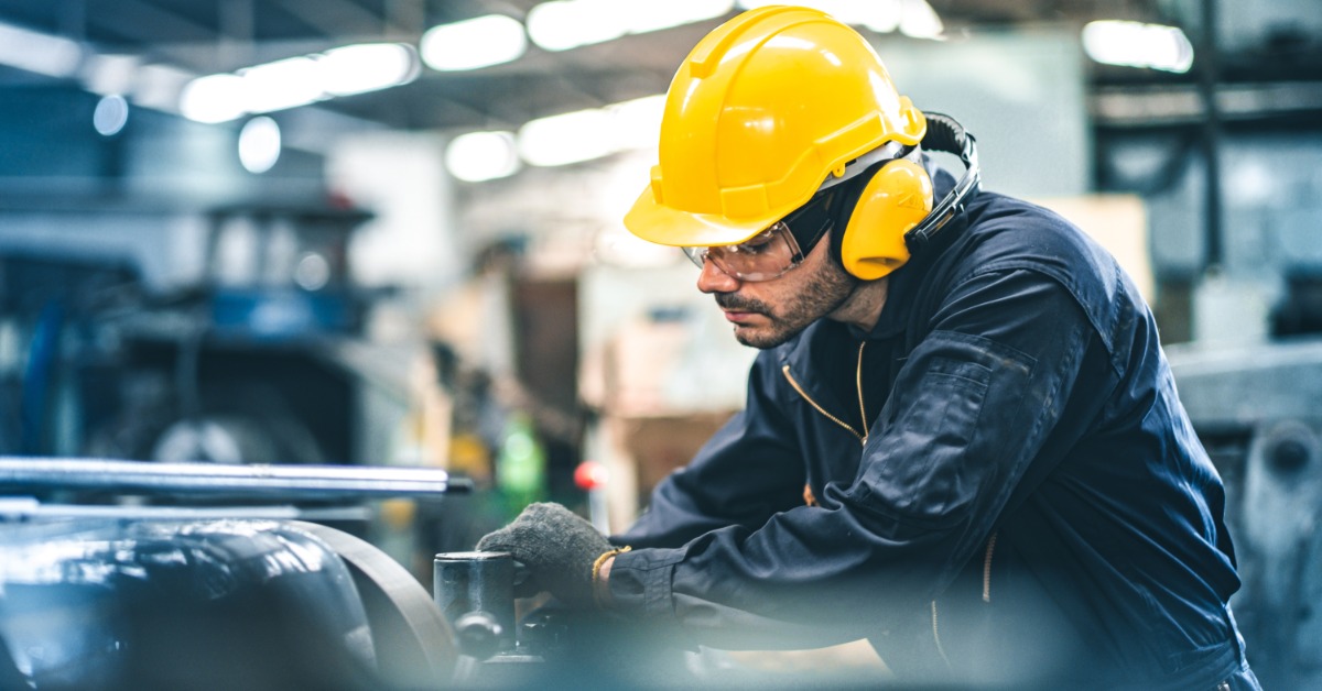 A man in a yellow hard hat is leaning over a piece of machinery as he works with it. The man is in a warehouse.