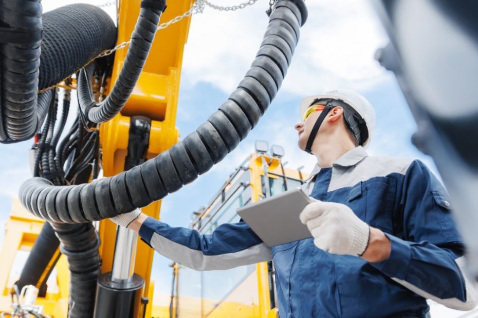 A man in a blue jumpsuit is standing next to a large piece of industrial equipment that is in an outdoor environment.