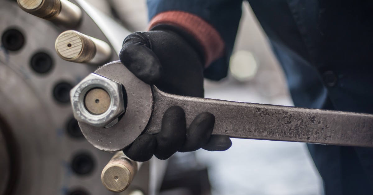 A person is using a large wrench on a bolt that is attached to a larger piece of metal. The man is wearing black gloves.