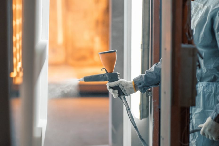 A person in a protective jumpsuit is spraying a chemical on a slab of metal. The metal is suspended from the ceiling.