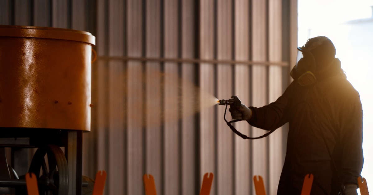 A person who is wearing a protective mask is using an applicator to apply a coating to a large piece of metal.