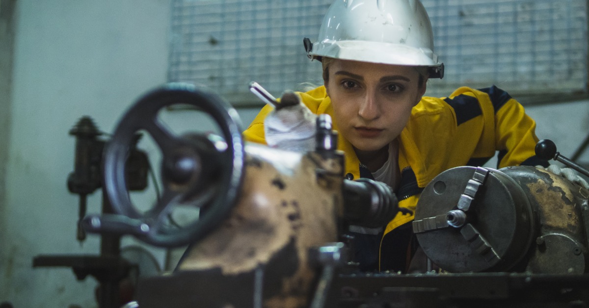 A woman is standing and looking at a piece of machinery that is sitting on a table. She is wearing a hard hat.