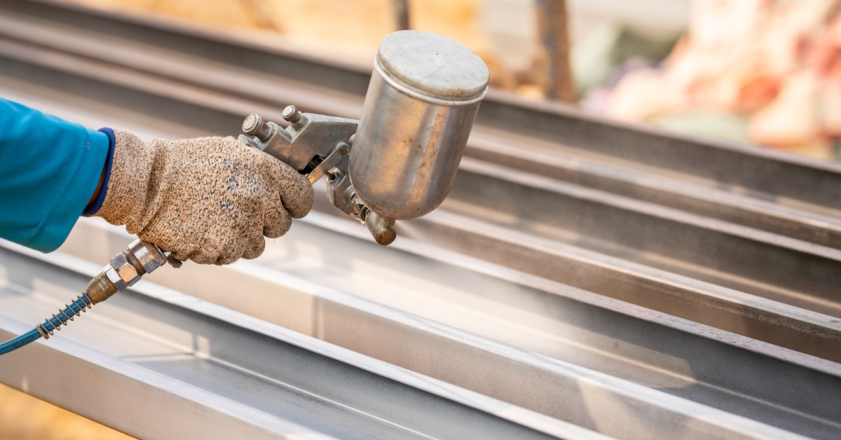 A person is holding a spray gun and is pointing it at several pieces of metal. The person is wearing a blue shirt.