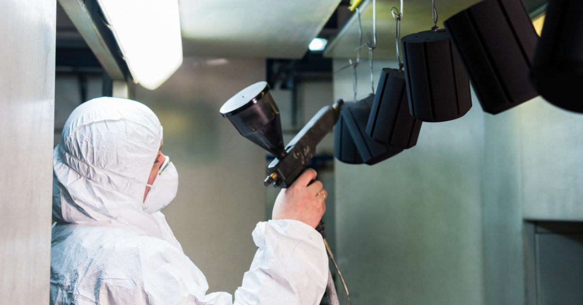 A person is wearing a white jumpsuit and is using a spray gun to spray some pieces of metal that are hanging from the ceiling.