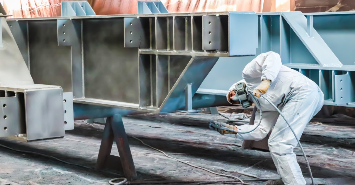 A person in a white bodysuit is kneeling next to a large metal object. They are spraying it with a spray gun.