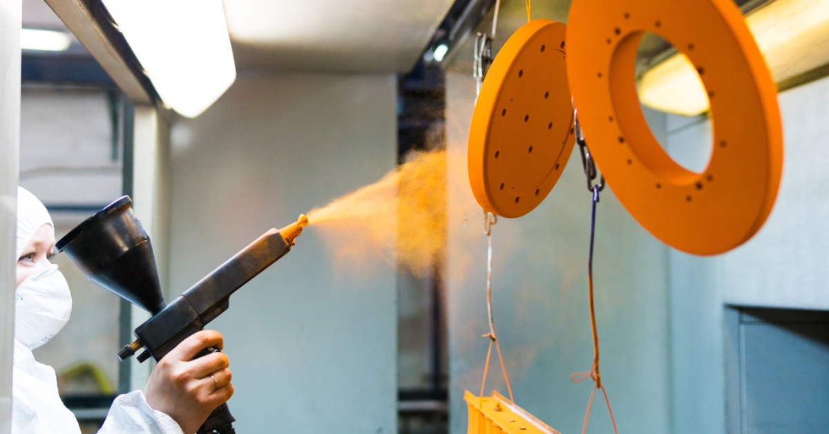 A woman in a face mask is using a spray gun to spray several pices of metal that are suspended from the ceiling.