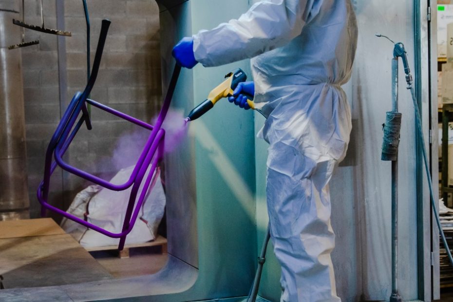 A person dressed in white protective clothing is spraying a suspended piece of metal in a factory setting.